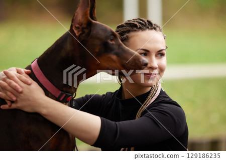Woman with braided hair hugging a Doberman outdoors in a park setting Woman with braided hair hugging a Doberman outdoors in a park setting 129186235