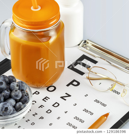 Glass of natural carrot juice, bowl with blueberries, vitamins, eyeglasses and Snellen table for vision testing. Nutritions for good vision. Vitamin A product to improve vision. Closeup 129186396