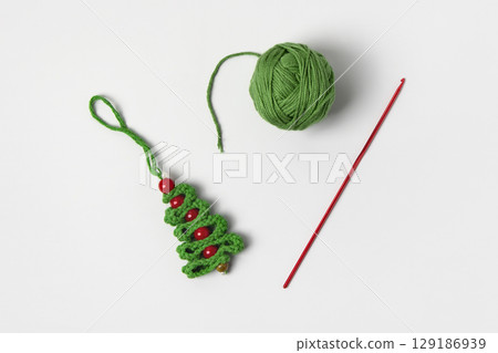 Crocheted green Christmas tree with red beads, a red knitting hook, and a green ball of yarn on a gray background. Home craft and knitting desk for Christmas toys and decorations 129186939