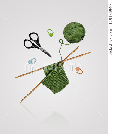 Tools for knitting warm clothes: scissors, green ball of thread, stoppers, and knitting needles with a knitted green piece of fabric flying in the air on a gray background. Hobby home crafts. 129186940