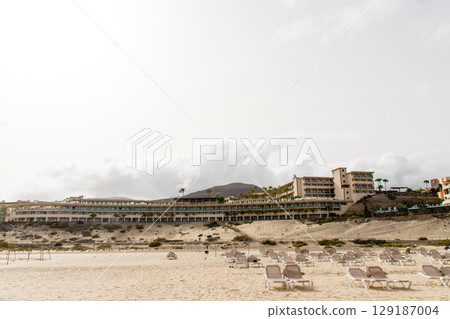 Beach beds lined up on a spacious sandy beach and a coastal resort hotel on Fuerteventura Island 129187004