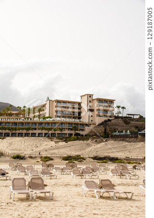 Beach beds lined up on a spacious sandy beach and a coastal resort hotel on Fuerteventura Island Beach beds lined up on a spacious sandy beach and a coastal resort hotel on Fuerteventura Island 129187005