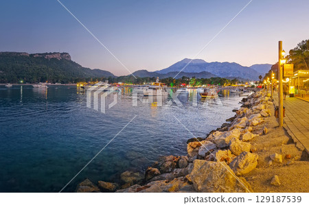 KEMER, TURKEY - JUNE 30, 2025 Moonlight beach in Kemer at dusk with boats illuminating the water. Lively atmosphere at the bay with beautiful Calis hill in the evening background KEMER, TURKEY - JUNE 30, 2025 Moonlight beach in Kemer at dusk with boats illuminating the water. Lively atmosphere at the bay with beautiful Calis hill in the evening background 129187539