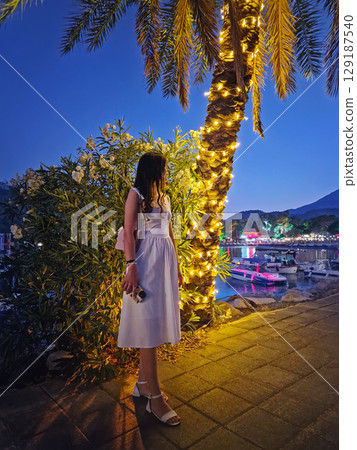 Charming evening scene with a gorgeous woman in a white dress, stands by a palm tree adorned with festive string lights, looking at the vibrant Moonlight beach at night in Kemer, Turkey Charming evening scene with a gorgeous woman in a white dress, stands by a palm tree adorned with festive string lights, looking at the vibrant Moonlight beach at night in Kemer, Turkey 129187540