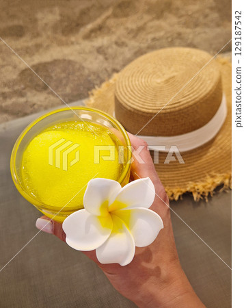 Young woman slender hand gently cradles a glass of vibrant yellow tropical drink, crowned by a delicate plumeria flower. Beach relaxation background with a straw sun hat on sand. Carefree summer vibes Young woman slender hand gently cradles a glass of vibrant yellow tropical drink, crowned by a delicate plumeria flower. Beach relaxation background with a straw sun hat on sand. Carefree summer vibes 129187542