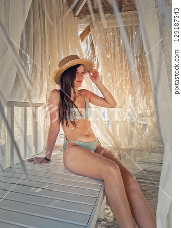 Beautiful young woman in pink bikini wearing a beach straw hat, relaxes on the lounge in a thatched beach hut with white curtains swaying gently in the sea breeze Beautiful young woman in pink bikini wearing a beach straw hat, relaxes on the lounge in a thatched beach hut with white curtains swaying gently in the sea breeze 129187543