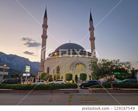 KEMER, TURKEY - JULY 1, 2025 Evening scene in Kemer featuring the grand mosque Huzur Camii Dzamija with two minarets and dome, and Taurus mountains at horizon. Active city life with street traffic KEMER, TURKEY - JULY 1, 2025 Evening scene in Kemer featuring the grand mosque Huzur Camii Dzamija with two minarets and dome, and Taurus mountains at horizon. Active city life with street traffic 129187549