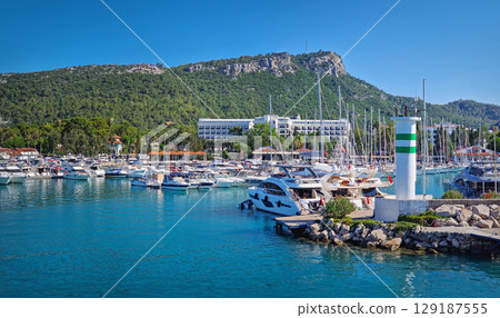 Kemer marina filled with numerous boats and yachts with Calis Hill in the background. A lighthouse stands prominently on the rocky pier, overlooking the clear blue water under a bright blue sky Kemer marina filled with numerous boats and yachts with Calis Hill in the background. A lighthouse stands prominently on the rocky pier, overlooking the clear blue water under a bright blue sky 129187555