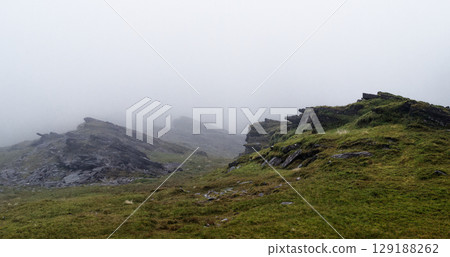 Rocky mountains covered with grass and shrubbery are almost completely shrouded in thick fog. The low visibility suggests a cloudy, overcast day in the Irish landscape. 129188262