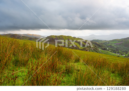 Grassy fields in the foreground lead to green, rocky hills under a cloudy sky in Coomloughra, Ireland. Mountain range visible in distance. Rural Irish landscape. 129188280
