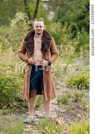 Muscular Man in Sheepskin Coat and Shorts Outdoors, Natural Environment, Green Foliage, Brown Fur, Bold Style, Rustic Setting, Eye-Catching Look 129188404