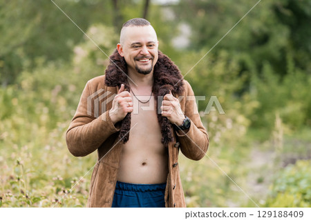 Handsome Man Outdoors in Brown Shearling Coat, Smiling, Open Jacket, Natural Scenery, Lush Green Forest Background, Warm Tone Photography. 129188409