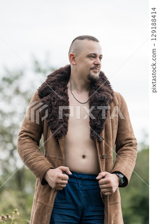 Portrait of a Rugged Man in a Brown Suede Coat with Fur Collar, Outdoors, Looking Away, Nature Background, Masculine Style, Cinematic Shot. Portrait of a Rugged Man in a Brown Suede Coat with Fur Collar, Outdoors, Looking Away, Nature Background, Masculine Style, Cinematic Shot. 129188414