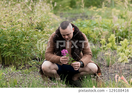 Man Kneeling in Meadow with Pink Flower, Nature Scene, Rustic Brown Fur Coat, Green Field, Soft Lighting, Emotional Moment, Springtime Pastoral. Man Kneeling in Meadow with Pink Flower, Nature Scene, Rustic Brown Fur Coat, Green Field, Soft Lighting, Emotional Moment, Springtime Pastoral. 129188416