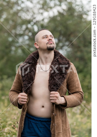 Rugged Man in Suede Coat Outdoors. Natural Forest Backdrop. Bare Chest. Earth Tones. Dramatic Lighting. Portrait of Strong Male Figure. Looking Up. Rugged Man in Suede Coat Outdoors. Natural Forest Backdrop. Bare Chest. Earth Tones. Dramatic Lighting. Portrait of Strong Male Figure. Looking Up. 129188423
