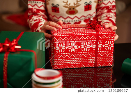Woman wearing Christmas sweater holding wrapped Christmas gift with wrapping paper and ribbon table. Preparing presents for winter holidays Woman wearing Christmas sweater holding wrapped Christmas gift with wrapping paper and ribbon table. Preparing presents for winter holidays 129189637