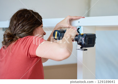 Woman using cordless drill to screw part into white bed frame. Female assembling furniture in new apartment. DIY home renovation and home organization after moving concept Woman using cordless drill to screw part into white bed frame. Female assembling furniture in new apartment. DIY home renovation and home organization after moving concept 129189641