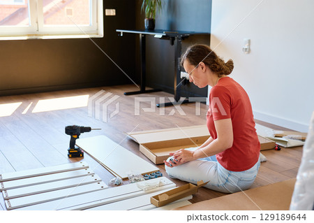 Woman unpacking parts for furniture assembly at home. Female sitting on floor and holding bed parts, wheels and hardware. Concept of DIY home improvement and renovation Woman unpacking parts for furniture assembly at home. Female sitting on floor and holding bed parts, wheels and hardware. Concept of DIY home improvement and renovation 129189644