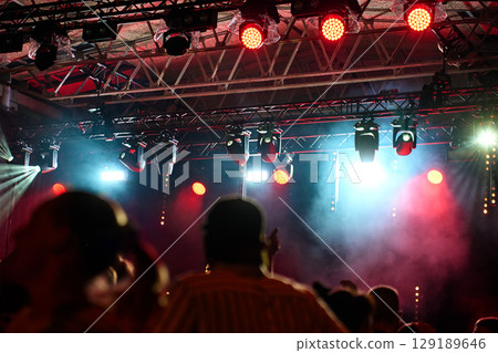 Crowd enjoying concert with bright lights and smoke on stage. Silhouettes of people dancing at night in front of concert stage with spotlights. Concept of live music event and entertainment Crowd enjoying concert with bright lights and smoke on stage. Silhouettes of people dancing at night in front of concert stage with spotlights. Concept of live music event and entertainment 129189646