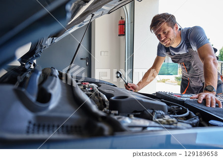 Male mechanic working on car engine with wrench under open hood in garage. Man in work overalls is repairing car engine with tool set in auto repair shop. Vehicle maintenance concept 129189658