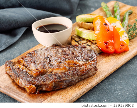 Fresh hot pork steak on wooden cutting board, served vegetables and sauce. Delicious pork steak on dark table. BBQ rib beefsteak or tender pork steak Fresh hot pork steak on wooden cutting board, served vegetables and sauce. Delicious pork steak on dark table. BBQ rib beefsteak or tender pork steak 129189808