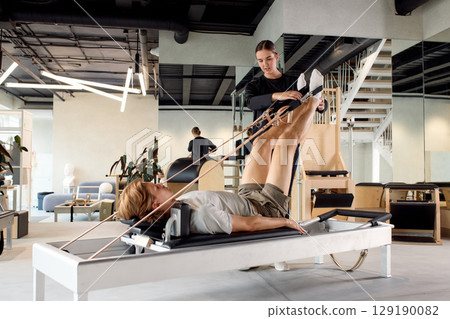 Trainer assisting a client with a rehabilitation pilates session in a modern studio Trainer assisting a client with a rehabilitation pilates session in a modern studio 129190082