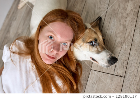 Red-haired woman and her dog lounging together on a wooden floor Red-haired woman and her dog lounging together on a wooden floor 129190117