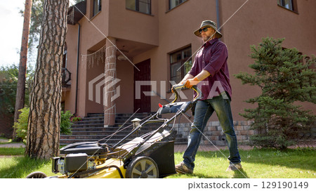 Man mowing the lawn in front of a house wearing a sun hat Man mowing the lawn in front of a house wearing a sun hat 129190149