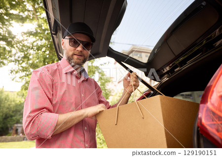 Man loading cardboard box into a car trunk outdoors on a sunny day 129190151