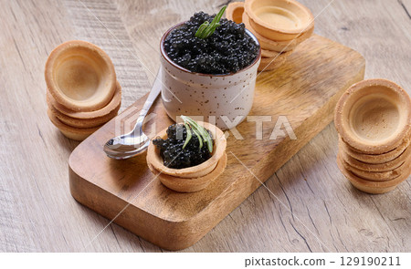 Black caviar, in a small ceramic pot and mini tartlet, placed on a wooden board. The composition includes a spoon and stacks of empty tartlets 129190211