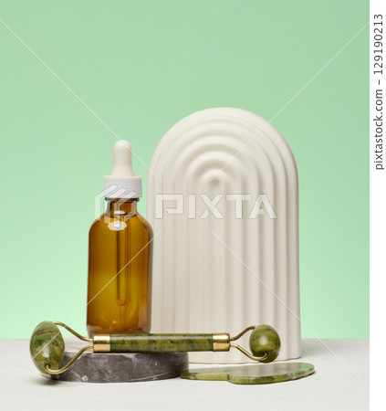 A bottle with a pipette, a jade facial massage roller and a decorative element are arranged on a green background. A bottle with a pipette, a jade facial massage roller and a decorative element are arranged on a green background. 129190213