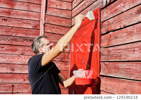 House owner applies paint to metal front door using flat paint brush, man in his fifties performing home maintenance task. House owner applies paint to metal front door using flat paint brush, man in his fifties performing home maintenance task. 129190218