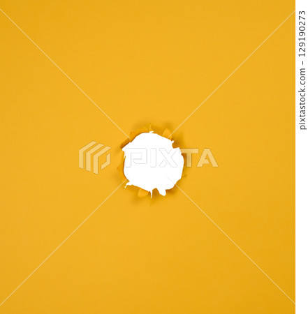 A round hole with jagged edges is made in a sheet of bright yellow paper. 129190273