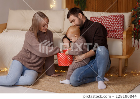 Happy young family with toddler playing on floor near sofa in cozy room with Christmas lights 129190844