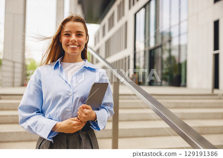 A confident woman stands outside a modern office building, holding a tablet and exuding positivity 129191386