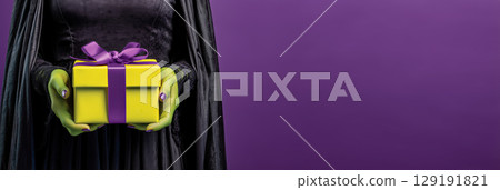 Person in black cloak holds yellow gift box with purple ribbon against purple background 129191821