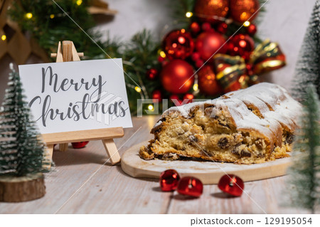 Note with saying MERRY CHRISTMAS congratulations. Traditional German Christmas stollen cake in background of handmade festive decor. Winter holiday treats. Vegan baking recipe 129195054