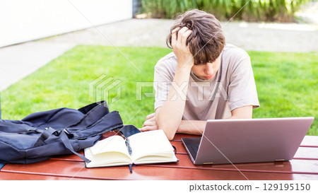 Tired of studying, a teenage boy sits with a laptop and a notebook, his head bowed in disappointment Tired of studying, a teenage boy sits with a laptop and a notebook, his head bowed in disappointment 129195150
