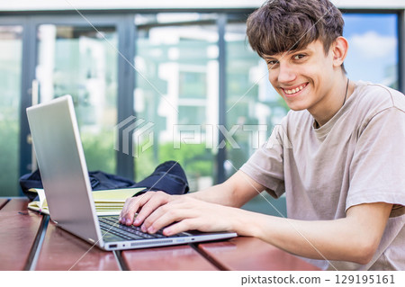 Cute happy teenager boy student with laptop at table outside in yard creating content and studying 129195161