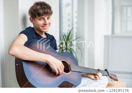 Cute cheerful teenage boy on balcony playing guitar, smiling at camera 129195176