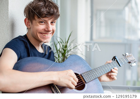 Cute cheerful teenage boy on balcony playing guitar, smiling at camera 129195179