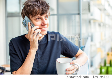 Cute teenage boy with a cup of coffee or tea happily talking on the phone on the balcony Cute teenage boy with a cup of coffee or tea happily talking on the phone on the balcony 129195182