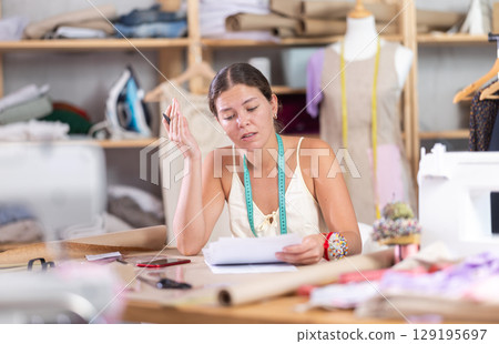 Young tailoress engaged in paperwork in sewing workshop 129195697