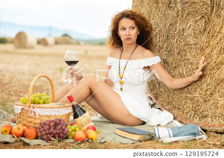 Woman in white dress sipping wine at rural picnic by hay bale Woman in white dress sipping wine at rural picnic by hay bale 129195724