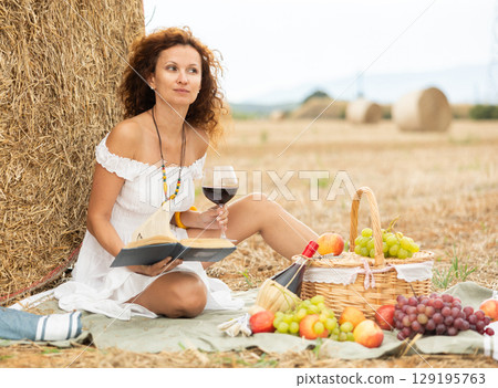 Woman sitting on ground in field, settled down in nature with book and glass of wine 129195763