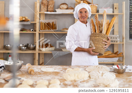 Positive senior woman baker in white uniform baking fresh baguettes in bakery 129195811