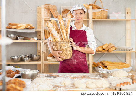 Smiling female baker holding wicker basket of golden baguettes Smiling female baker holding wicker basket of golden baguettes 129195834