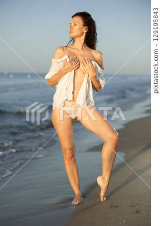 Sexy woman in white lingerie and mesh cape walking along the seashore in surf at sunset 129195843