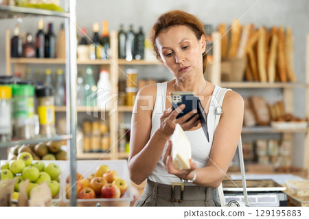 Female shopper scans QR code on cheese label using smartphone. Checking products in supermarket 129195883