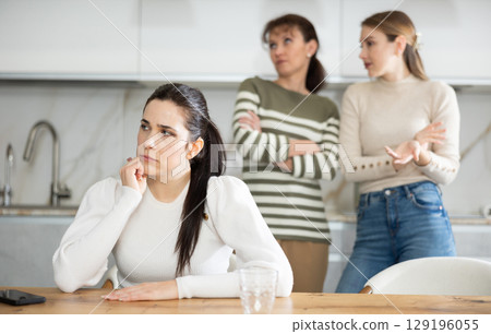 Upset middle-aged woman sitting at the kitchen table with her back to women quarreling to her Upset middle-aged woman sitting at the kitchen table with her back to women quarreling to her 129196055